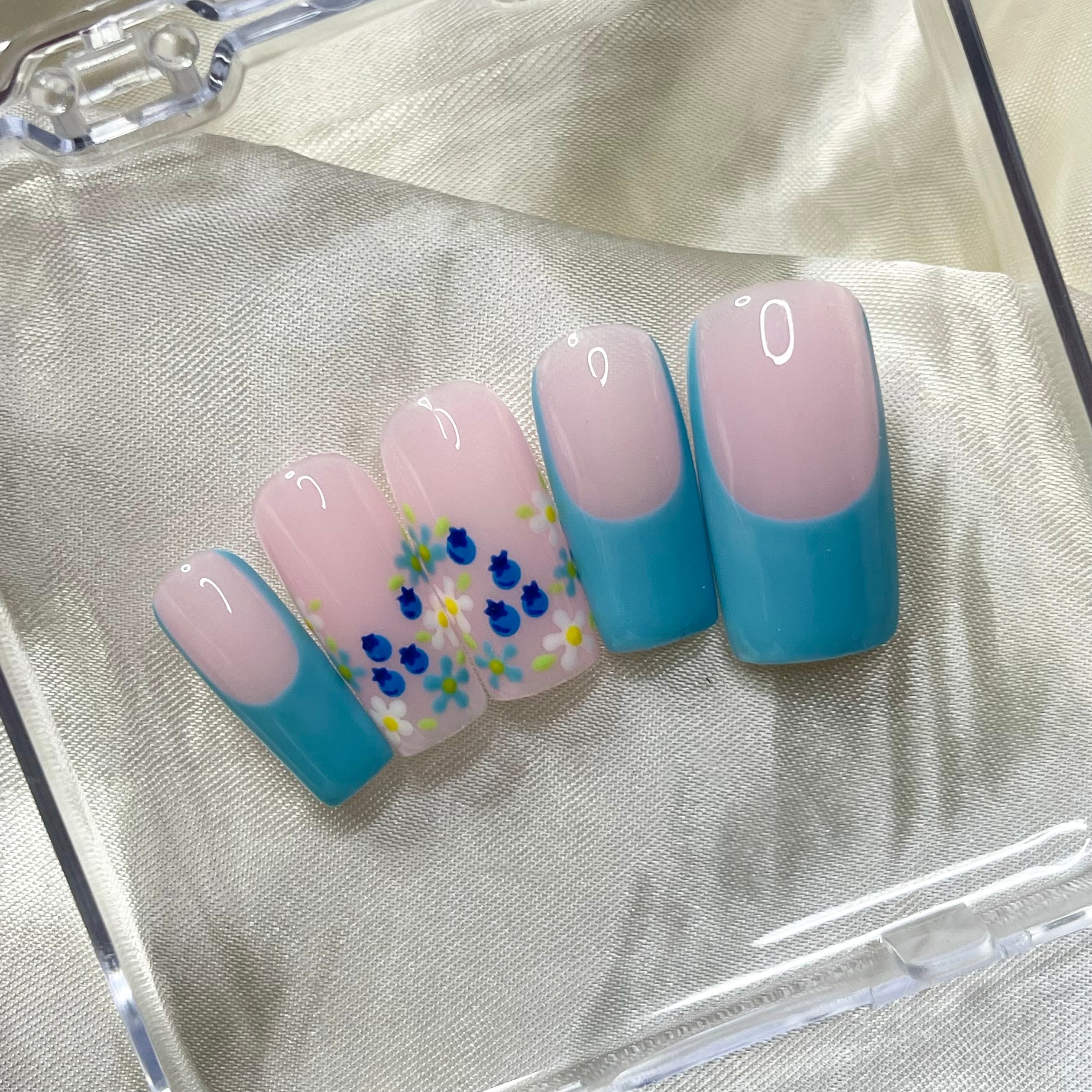 Blueberry Fields press-on nails feature blue French tips, floral designs, hand-painted blueberries, and soft pinky nude bases for a sweet, botanical spring look.
