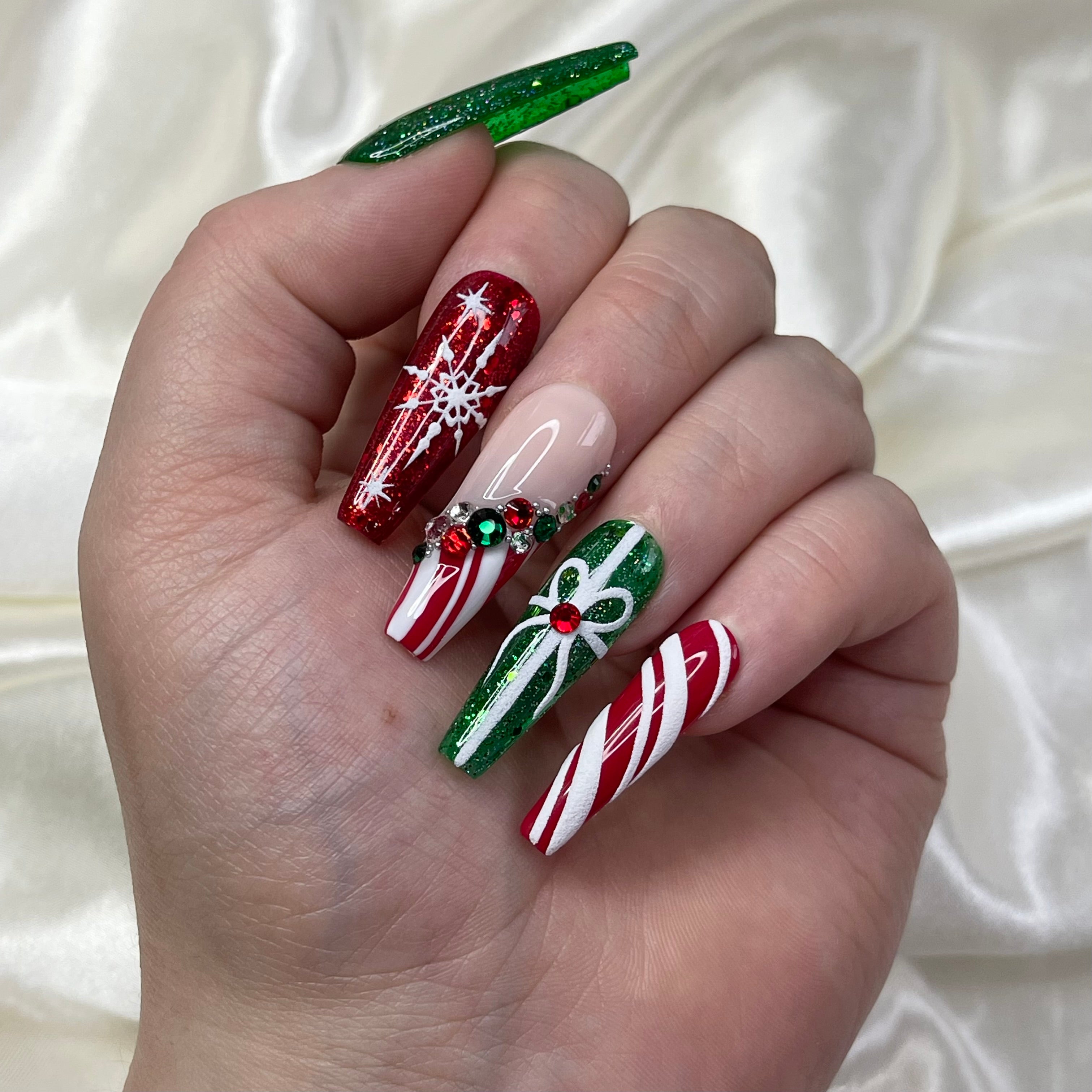 A set of artificial nails with a holiday theme, featuring green glitter, red glitter with snowflakes, and candy cane tips with crystals, all painted on a hand.