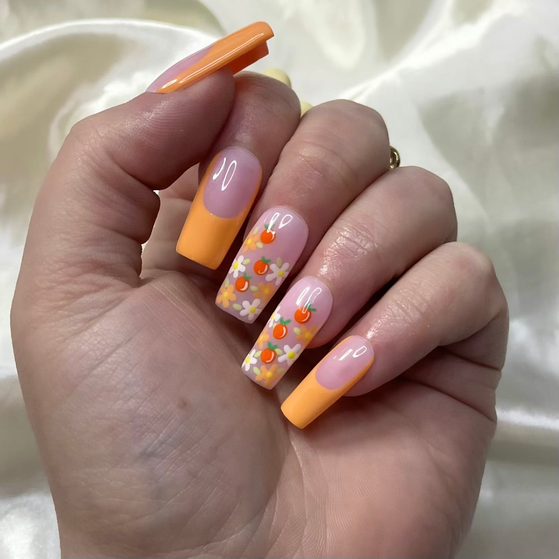 Tangerine Trails press-on nails feature bold orange French tips, white and orange flowers, golden centres, hand-painted oranges, and soft pinky nude bases for a zesty spring look.