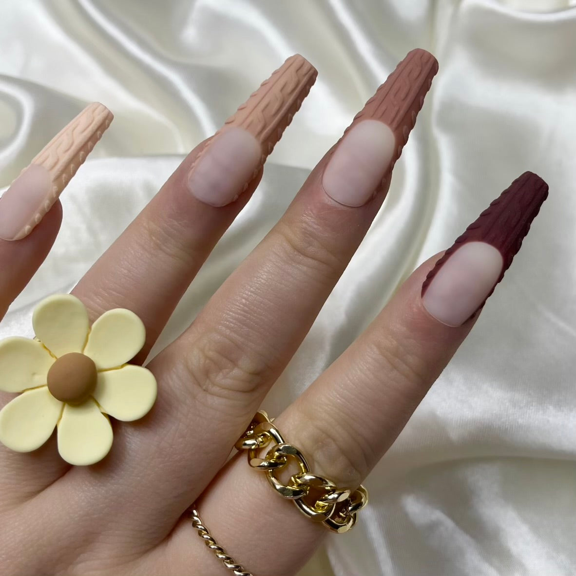 A manicure image showing matte nude French tips with a sweater-knit design on the nails, alongside a yellow ring with a floral pattern on the finger.