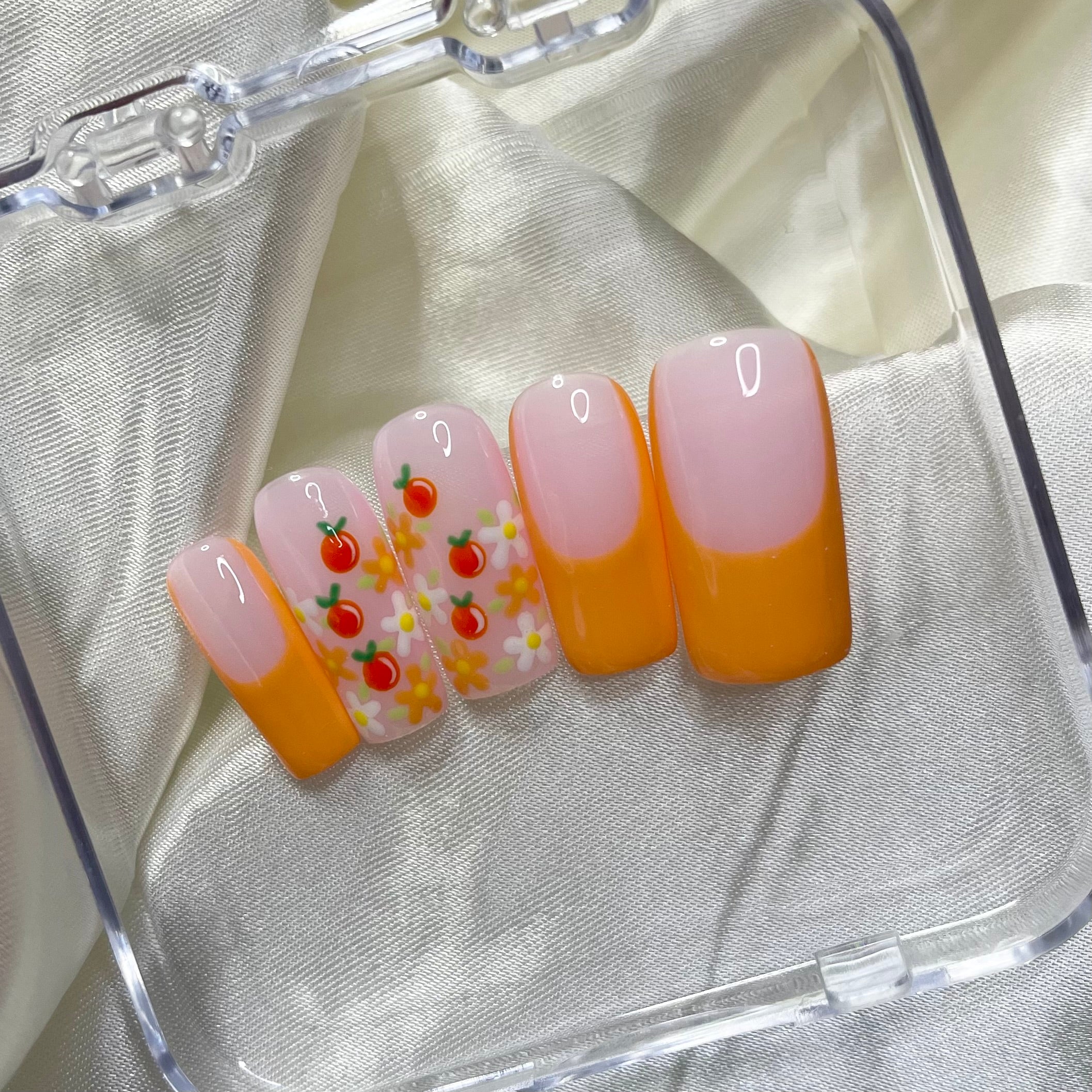 Tangerine Trails press-on nails feature bold orange French tips, white and orange flowers, golden centres, hand-painted oranges, and soft pinky nude bases for a zesty spring look.
