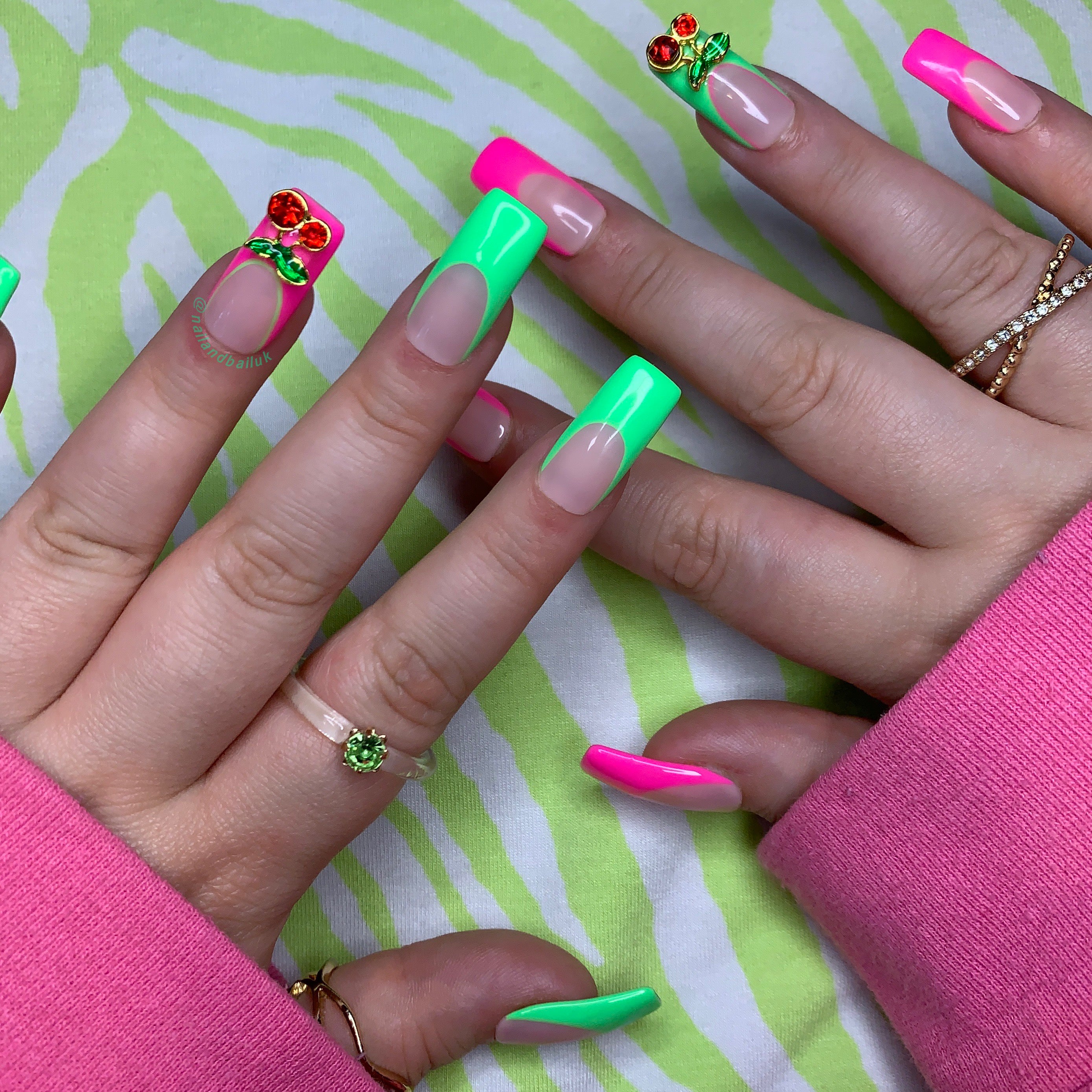 Nude press-on nails with neon pink and green French tips and cherry nail charms