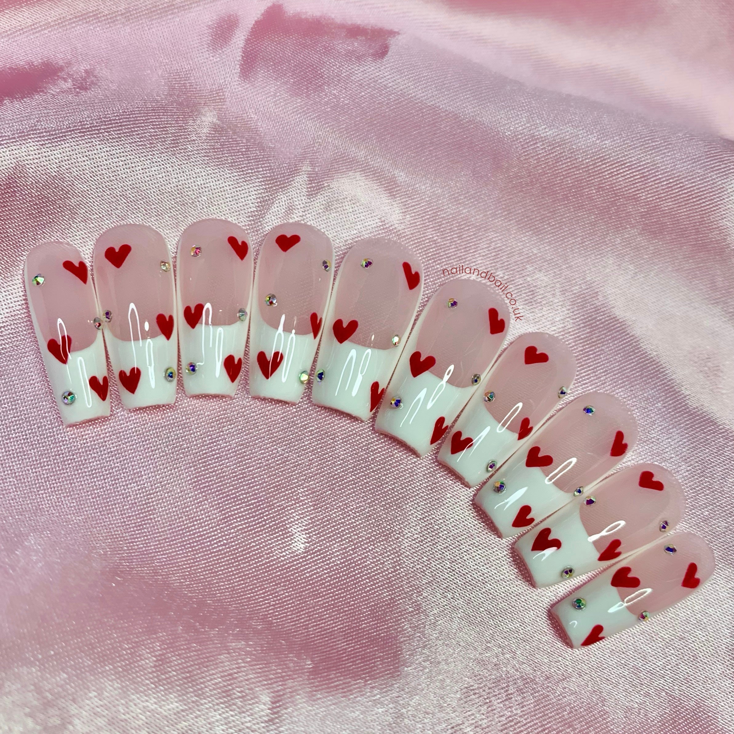 Nude press-on nails with white French tips, red heart nail art, and silver crystal details