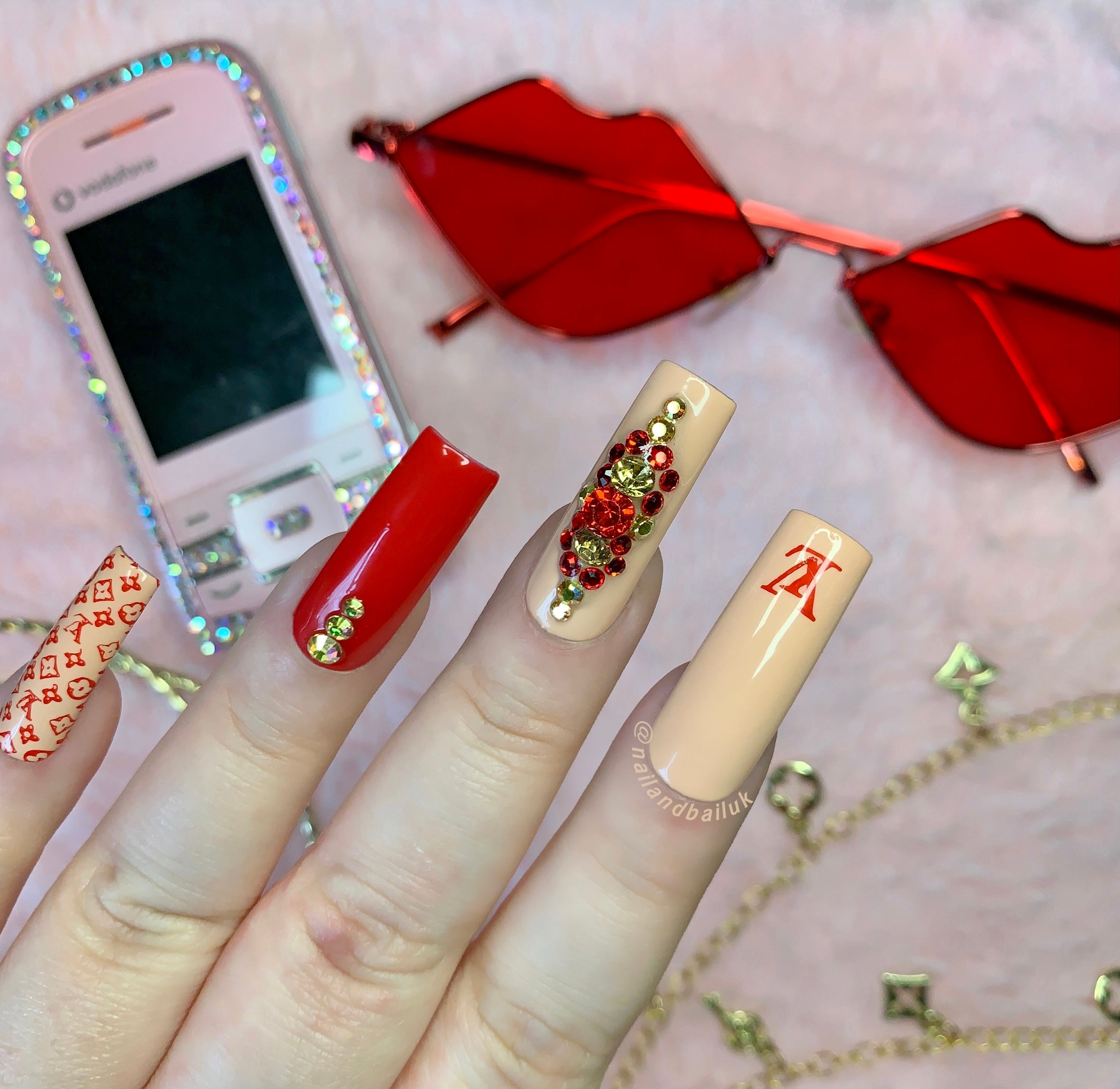 Nude and red press-on nails with luxury-inspired prints and red and gold crystal clusters